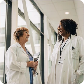 Two female doctors in lab coats smiling and chatting in a hospital hallway.