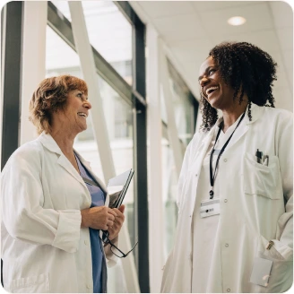 Two female doctors in lab coats smiling and chatting in a hospital hallway.