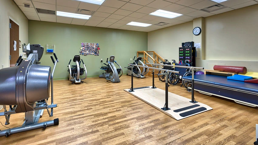 A physical therapy room with exercise equipment, parallel bars, and rehabilitation tools on hardwood floors.