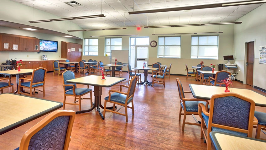 A spacious dining area with neatly arranged tables and chairs, a TV, and bright windows on wooden floors.