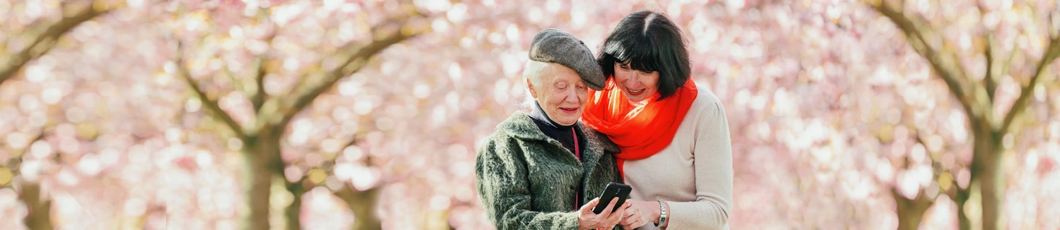 Two women, one elderly, stand under blooming trees, smiling while looking at a smartphone together.