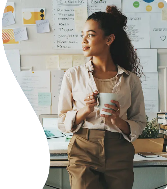 HC20731-senhea-cws-header-584x660px-prodev A woman stands in front of a desk holding a coffee mug, looking thoughtfully to the side; she is surrounded by notes, charts, and planning materials pinned to the wall behind her, suggesting a creative or project-focused workspace.