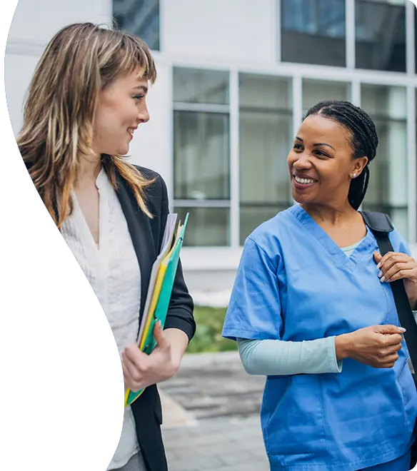 A smiling healthcare professional in blue scrubs walks and chats with a woman in business attire holding folders, both appearing engaged and friendly outside a modern office or hospital building.