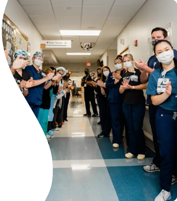 Hospital staff in scrubs and face masks form a corridor in a brightly lit hallway, clapping and smiling in what appears to be a celebratory or welcoming moment.