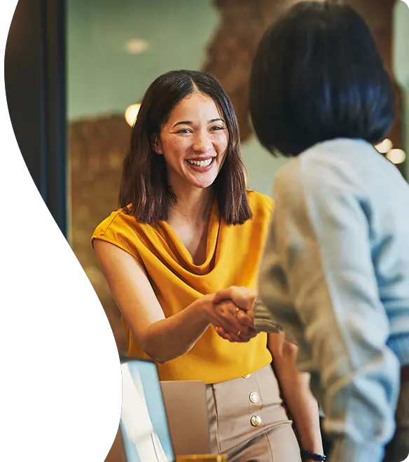 A cheerful woman in a mustard-yellow top smiles while shaking hands with another person in an indoor office setting, suggesting a friendly greeting or successful meeting.