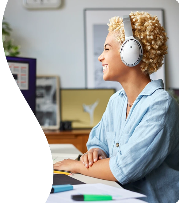 A cheerful young woman wearing headphones sits at a desk, smiling as she looks at a computer screen, surrounded by office supplies in a bright and creative workspace.