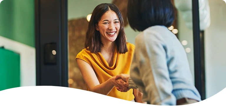 A cheerful woman in a mustard-yellow top smiles while shaking hands with another person in an indoor office setting, suggesting a friendly greeting or successful meeting.