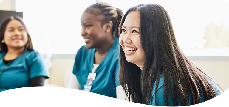 Smiling healthcare workers wearing teal-colored scrubs sit together in a bright room