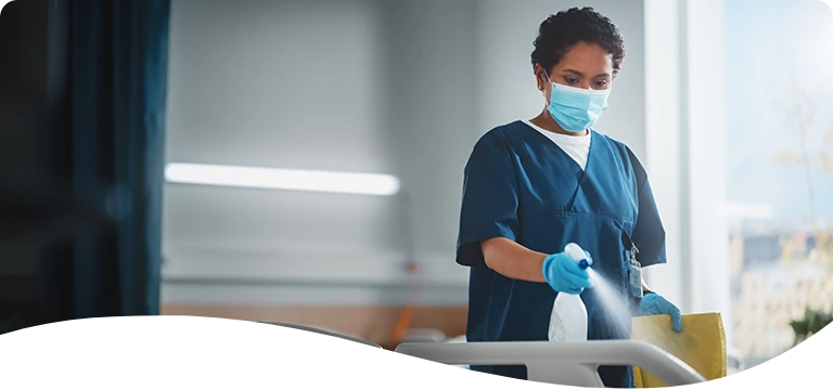 A healthcare worker wearing scrubs, gloves, and a face mask disinfects a hospital bed with a spray bottle and cleaning cloth in a brightly lit medical facility.