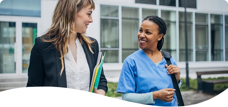 A smiling healthcare professional in blue scrubs walks and chats with a woman in business attire holding folders, both appearing engaged and friendly outside a modern office or hospital building.