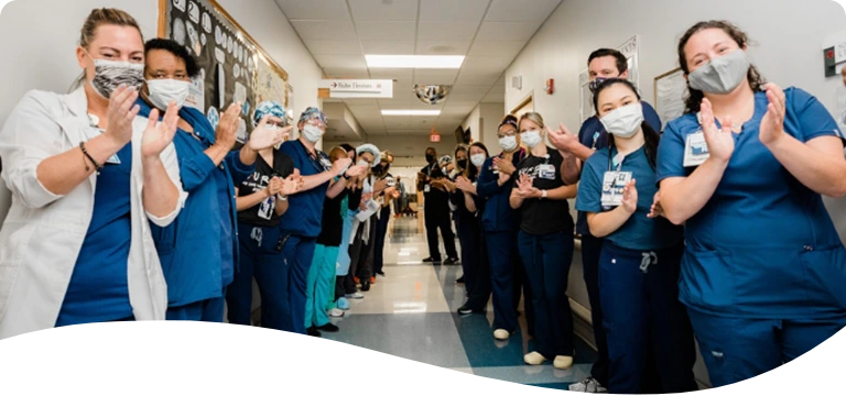 Hospital staff in scrubs and face masks form a corridor in a brightly lit hallway, clapping and smiling in what appears to be a celebratory or welcoming moment.