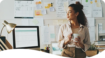 A woman stands in front of a desk holding a coffee mug, looking thoughtfully to the side; she is surrounded by notes, charts, and planning materials pinned to the wall behind her, suggesting a creative or project-focused workspace.