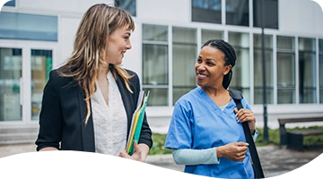 A smiling healthcare professional in blue scrubs walks and chats with a woman in business attire holding folders, both appearing engaged and friendly outside a modern office or hospital building.
