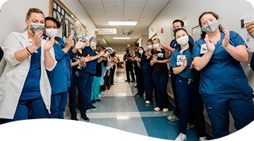 Hospital staff in scrubs and face masks form a corridor in a brightly lit hallway, clapping and smiling in what appears to be a celebratory or welcoming moment.