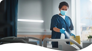 A healthcare worker wearing scrubs, gloves, and a face mask disinfects a hospital bed with a spray bottle and cleaning cloth in a brightly lit medical facility.