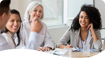 A group of professionals, including women in business attire and medical uniforms, sit around a conference table engaged in a cheerful and collaborative meeting in a bright, modern office setting.