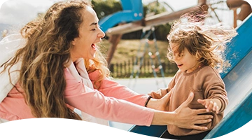 A joyful woman greets a laughing child at the bottom of a playground slide on a sunny day, both sharing a moment of happiness and connection.