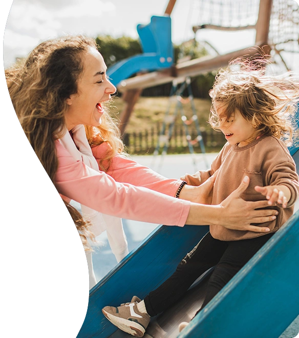 A joyful woman greets a laughing child at the bottom of a playground slide on a sunny day, both sharing a moment of happiness and connection.