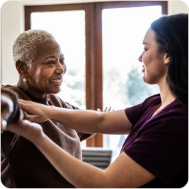 A woman smiling as a healthcare worker helps her with arm exercises in a bright, sunlit room.