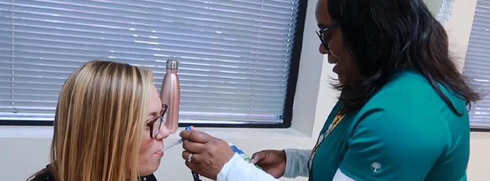 A healthcare worker in scrubs checks the temperature of a seated woman using a forehead thermometer.