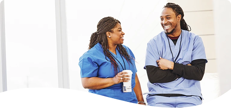 Two healthcare professionals in blue scrubs share a lighthearted conversation and smiles during a break, with one holding a coffee cup in a casual, relaxed setting.