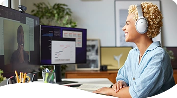 A cheerful young woman wearing headphones sits at a desk, smiling as she looks at a computer screen, surrounded by office supplies in a bright and creative workspace.