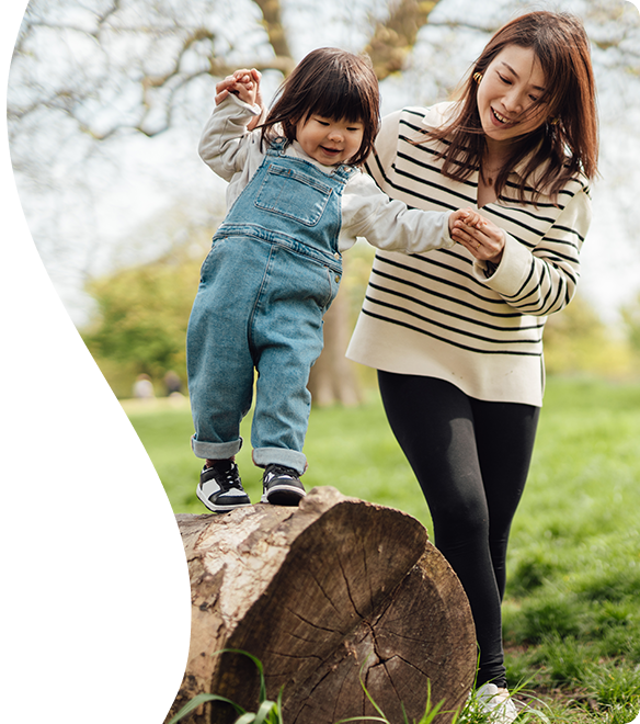 A woman helping a female toddler balance on a fallen tree outdoor on a sunny day.