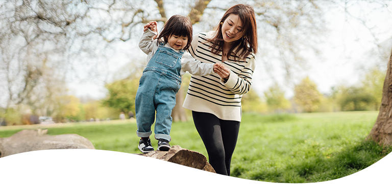 A woman helping a female toddler balance on a fallen tree outdoor on a sunny day.