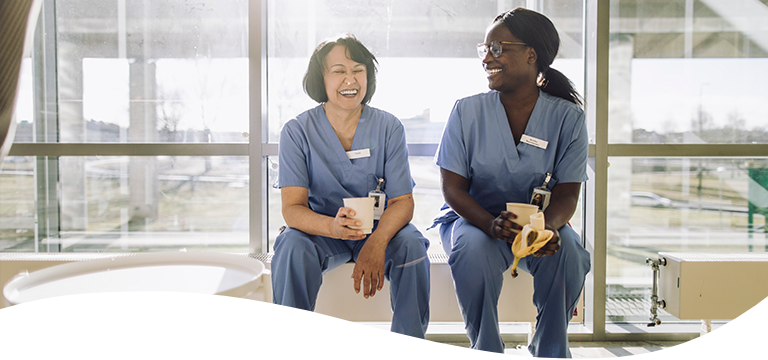 Two female nurses in scrubs laughing and enjoying a break with coffee and a banana in a sunshine filled office with glass wall behind them.