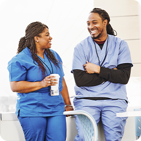 Two nurses in scrubs smiling and chatting during a break, one holding a coffee cup in a bright hospital setting.