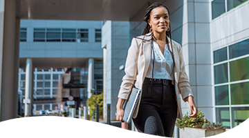 A woman dressed in business attire walking outside an office building with a laptop in hand.