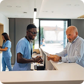 Smiling healthcare worker assists a male patient at a clinic front desk while another nurse works in the background.