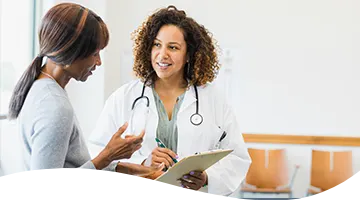 A smiling healthcare worker with a stethoscope around her neck taking notes on a clipboard while talking to another person.