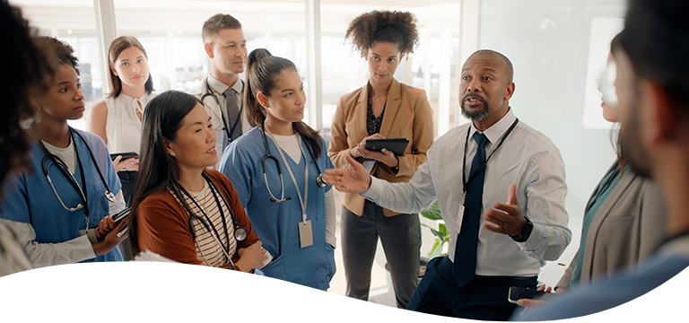 STUD360-senhea-cws-tablet-header-768x360px-supportservices (1) A man in business attire speaks to a group of healthcare professionals, some in scrubs and others in business clothing, gathered closely in a bright office setting.