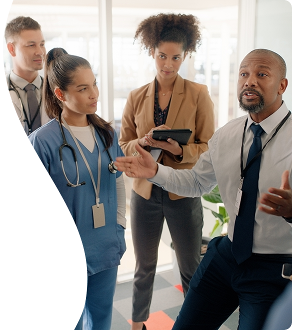 STUD360-senhea-cws-desktop-header-584x660px-supportservices (1) A man in business attire speaks to a group of healthcare professionals, some in scrubs and others in business clothing, gathered closely in a bright office setting.